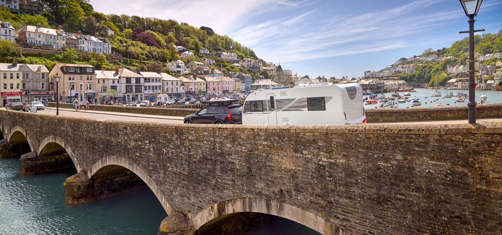  A family caravan is towed by a car over a scenic arched bridge, with the ocean in the background.
