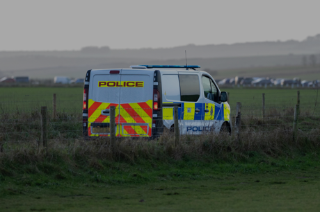 A police van parked in a field behind a wire fence, with a row of vehicles visible in the distance.
