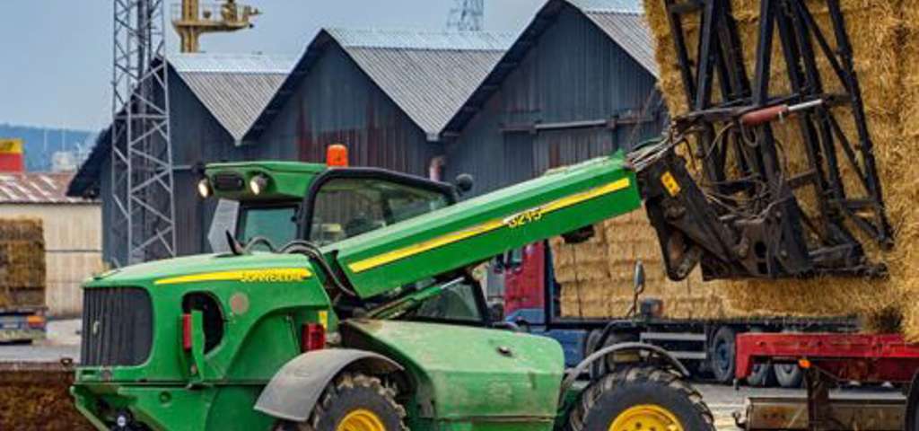 Green telehandler in action, unloading straw bales from a red lorry on a rural site.