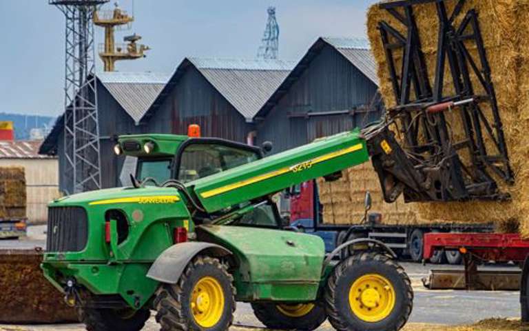 Green telehandler in action, unloading straw bales from a red lorry on a rural site.