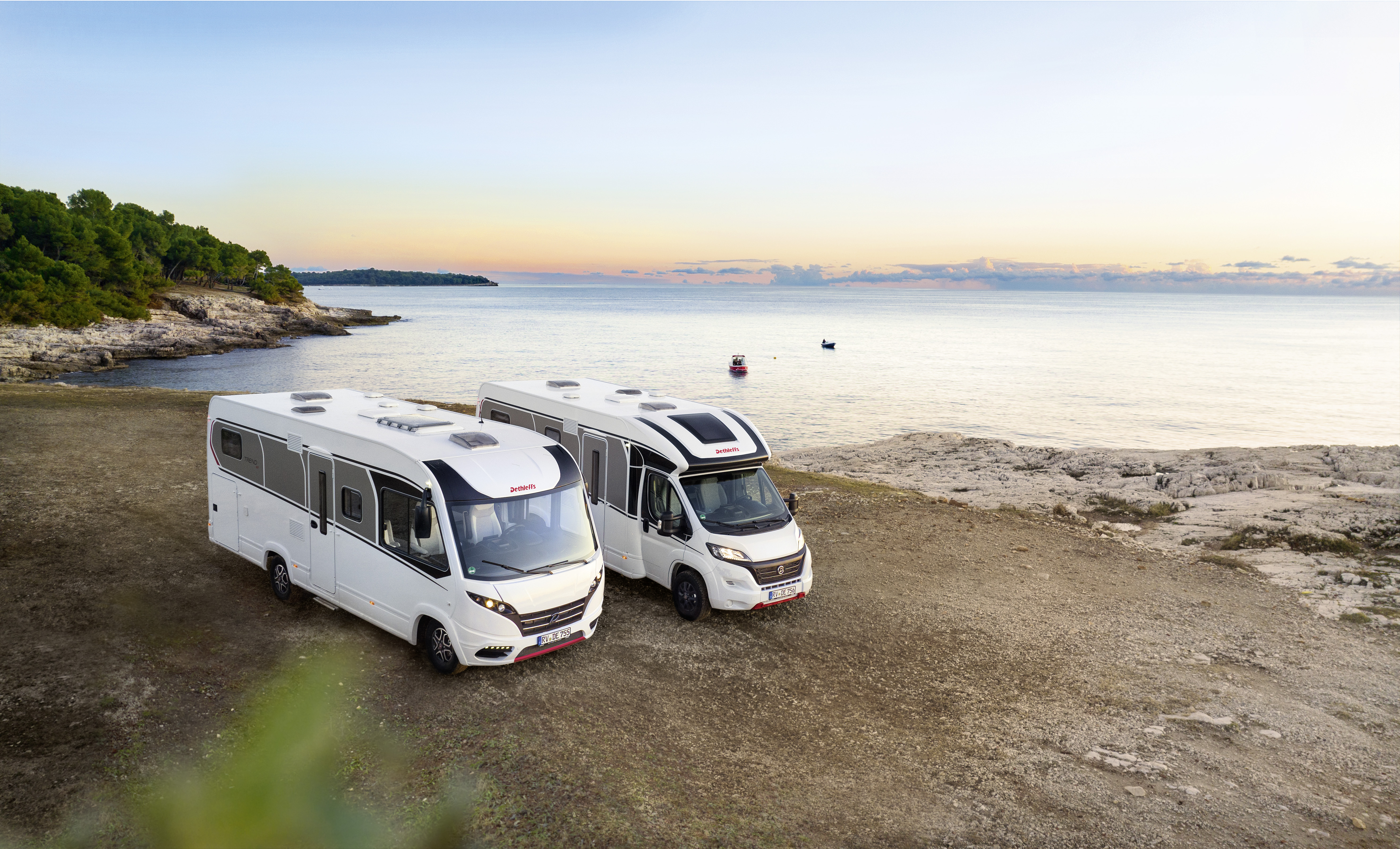 Two motorhomes parked near a coastal view as the sun sets over the horizon.