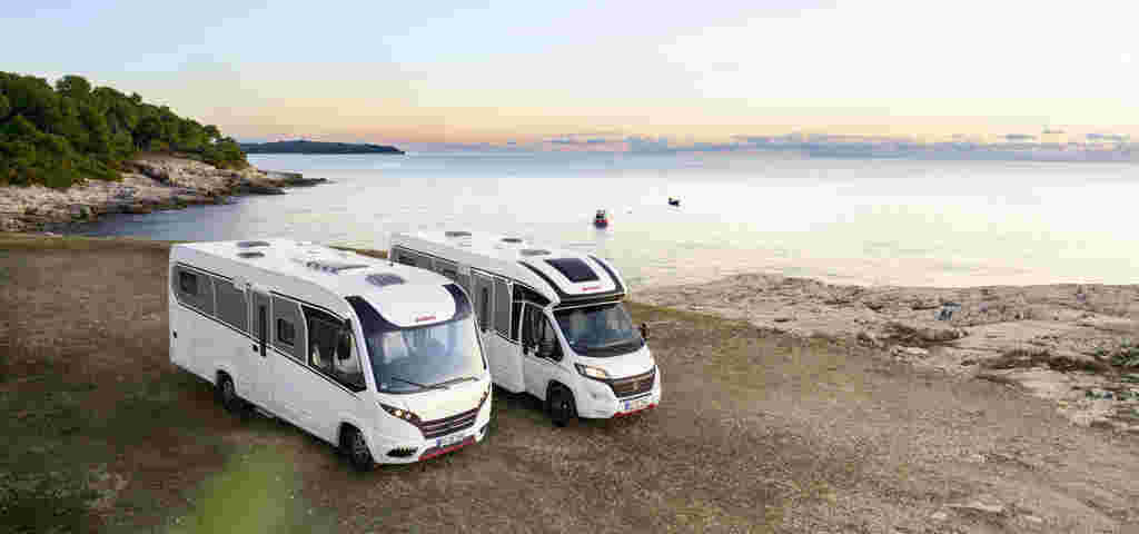 Two motorhomes parked near a coastal view as the sun sets over the horizon.
