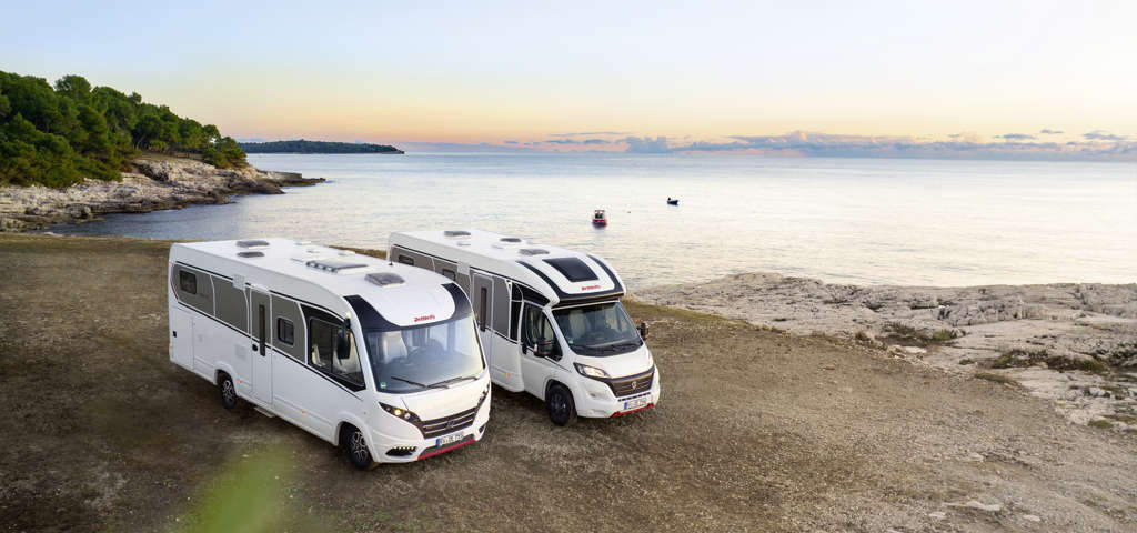 Two motorhomes parked near a coastal view as the sun sets over the horizon.