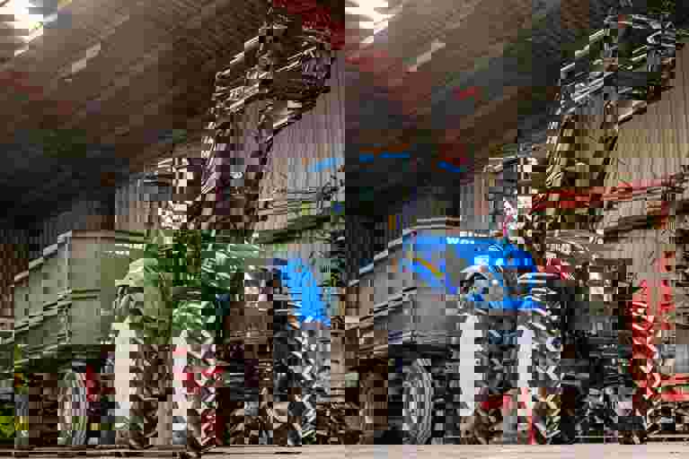 A blue utility loader tractor attached to a green trailer parked in a farmyard setting.