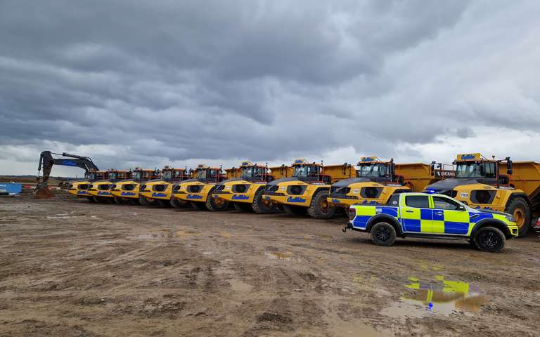 A police car parked to the right of a line of yellow off-road dumpers on a construction site.