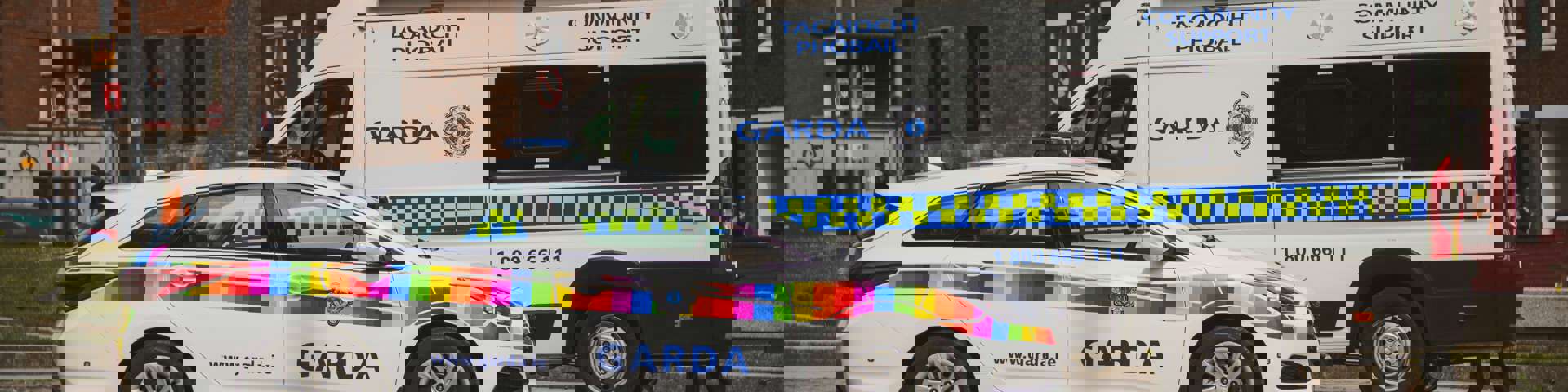 Rainbow flag-branded Garda car and a Garda community support van parked on a cobblestone street.