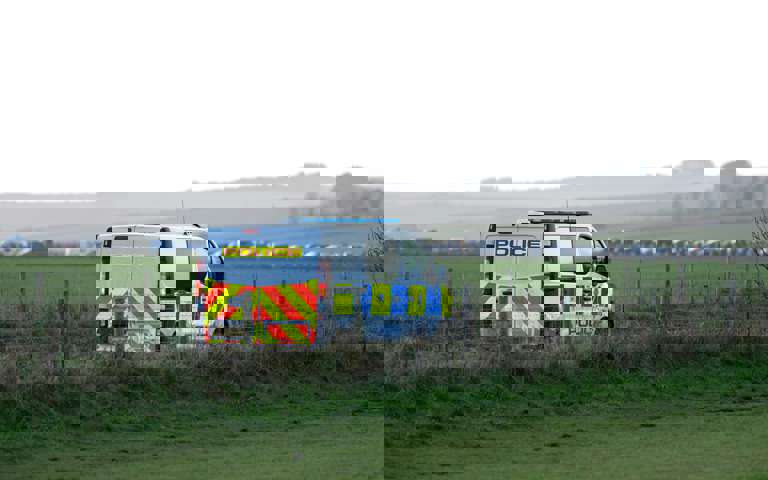 A police van parked in a field behind a wire fence, with a row of vehicles visible in the distance.