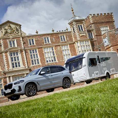 Silver car with a white caravan, stationary in front of an elegant stately home.