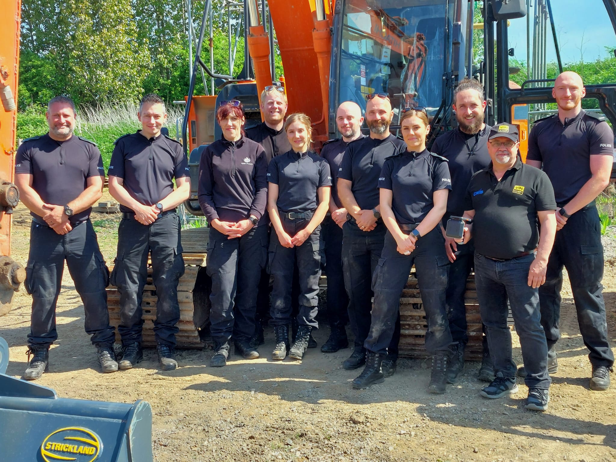 Ten Kent Police officers in blue uniforms with VIN Chip trainer, lined up in front of heavy machinery during IMI theft awareness training.