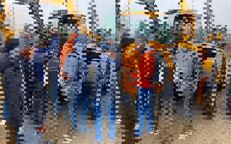 A group of students engaged in training, with JCBs and a rural landscape in the background.