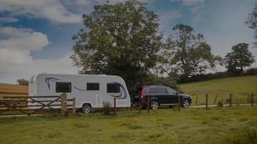 A black car towing a white caravan along a scenic route in vibrant green countryside.