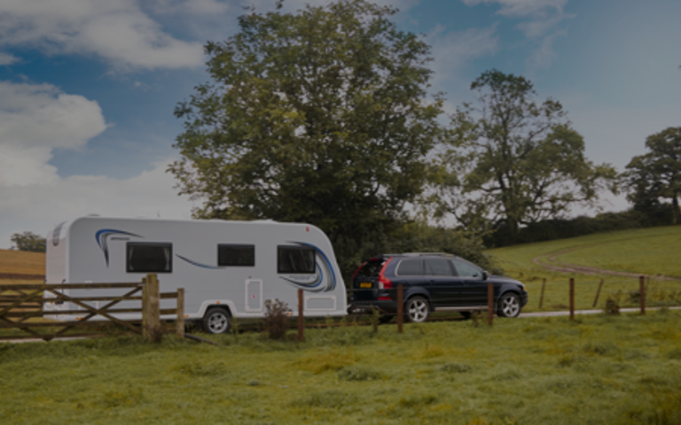 A black car towing a white caravan along a scenic route in vibrant green countryside.