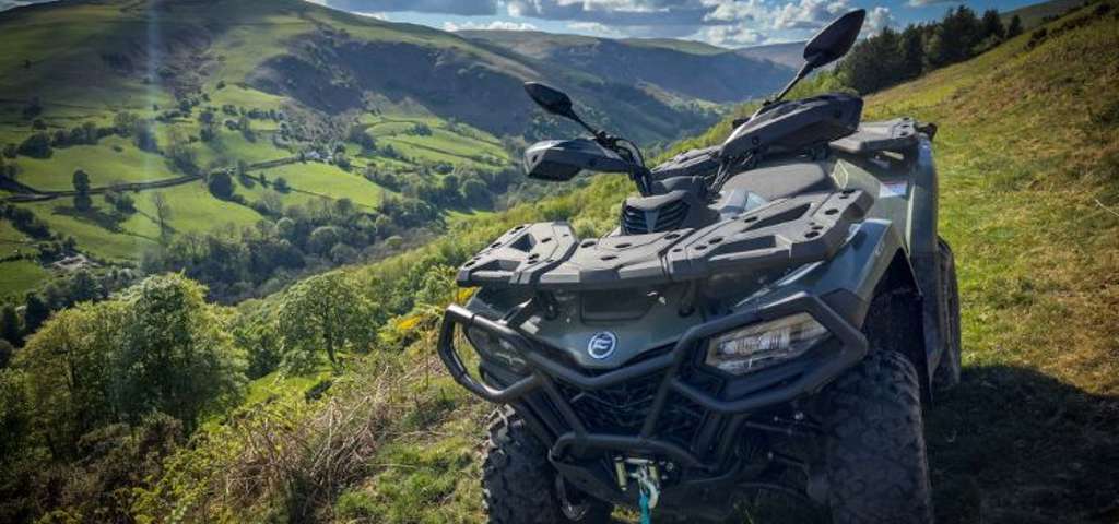 Green CFMOTO ATV parked on rolling hills under dramatic cloudy sky with sunbeams.
