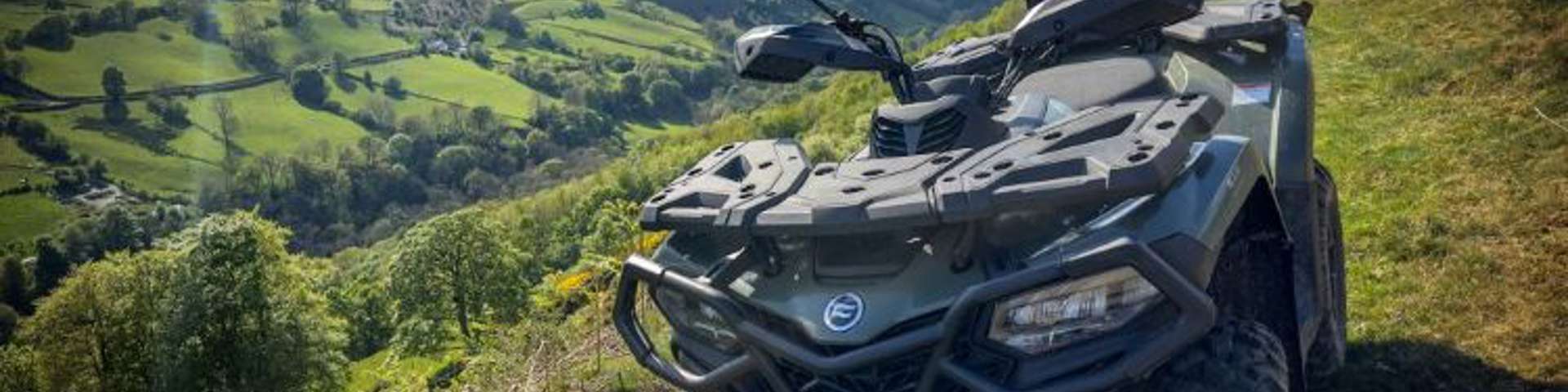 Green CFMOTO ATV parked on rolling hills under dramatic cloudy sky with sunbeams.