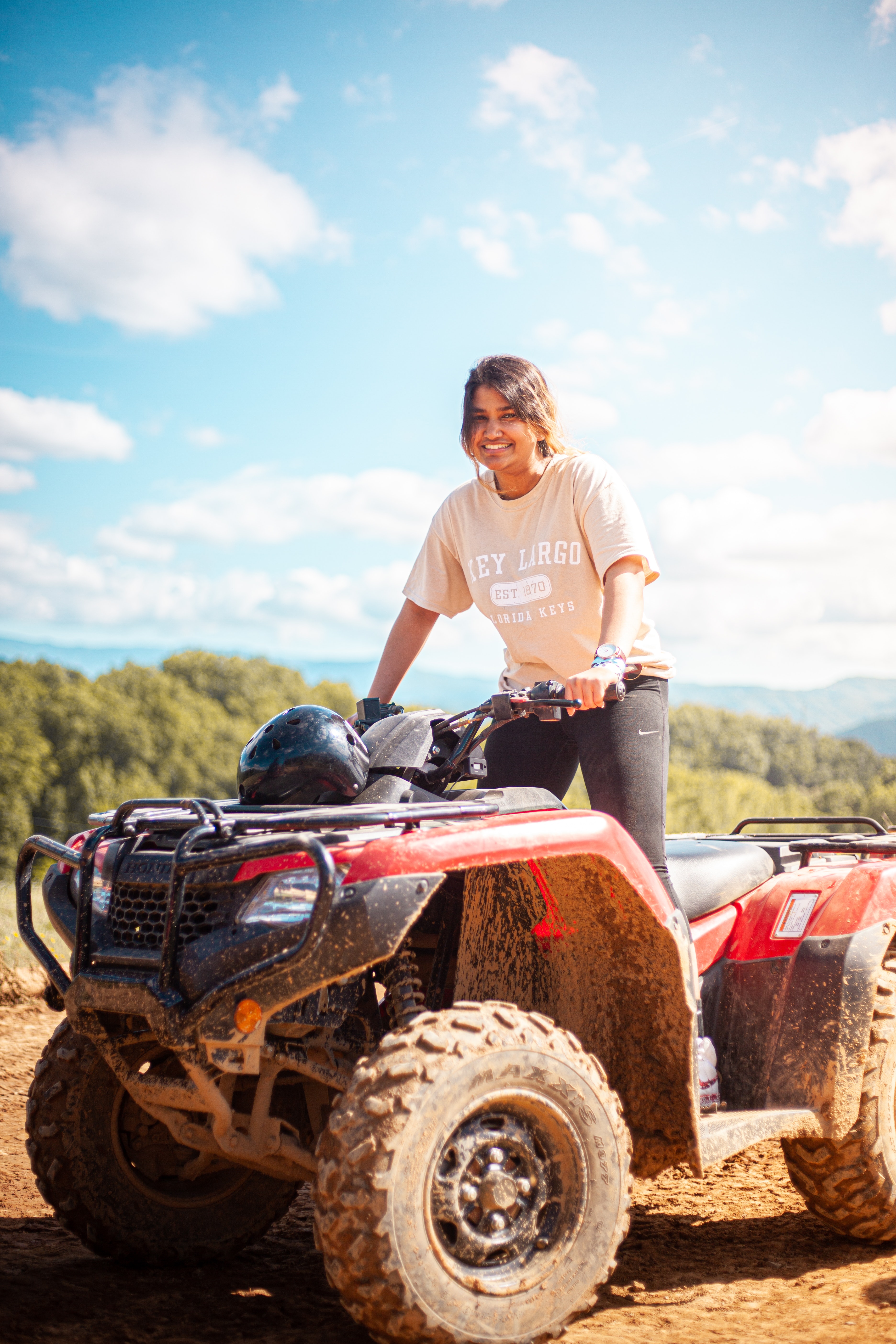 Female rider on a red quad bike, stationary against a backdrop of green trees and a partly cloudy blue sky.