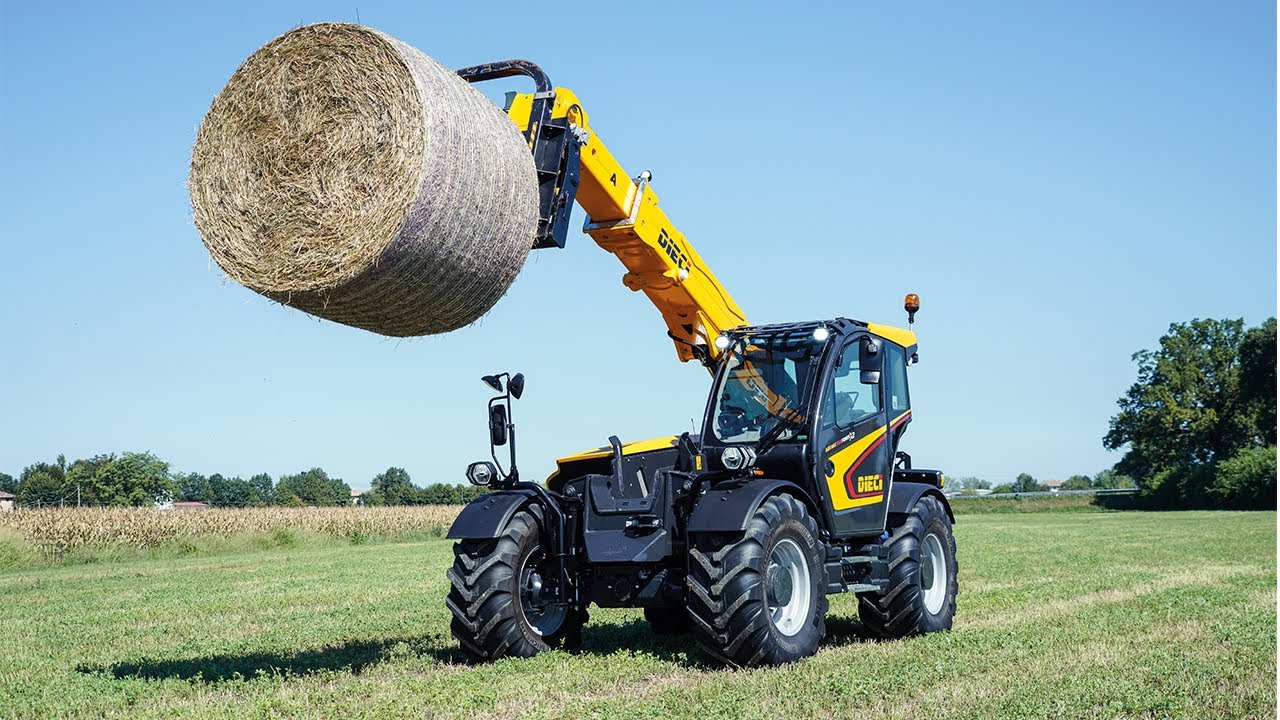 A yellow and black telehandler is lifting a round bale in an open, green field.