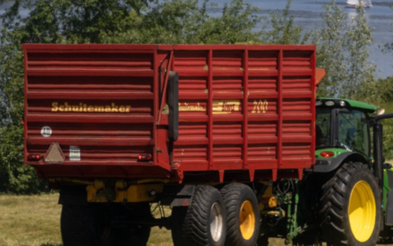 A green tractor pulls a bright red-orange agricultural trailer in a field by a lake, with a sailboat visible in the distance.