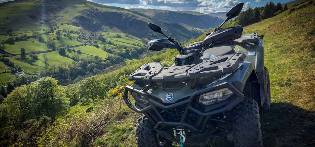 Green CFMOTO ATV parked on rolling hills under dramatic cloudy sky with sunbeams.