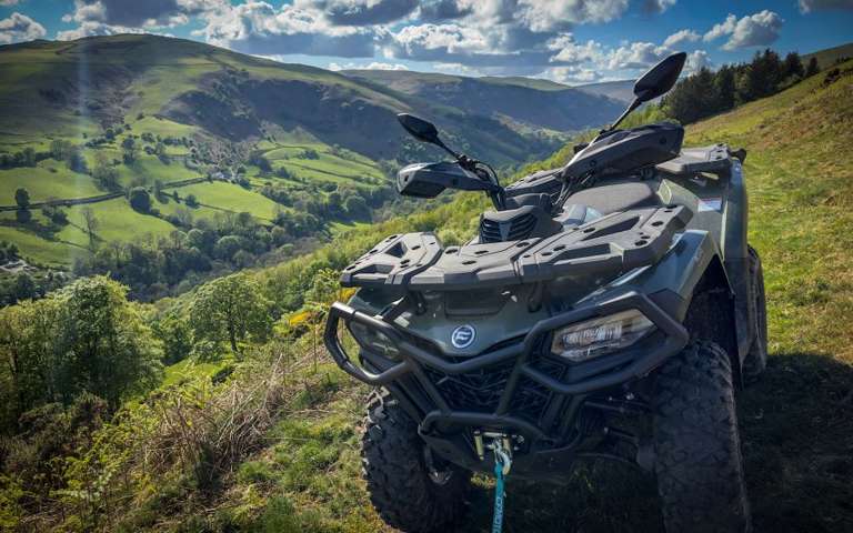 Green CFMOTO ATV parked on rolling hills under dramatic cloudy sky with sunbeams.