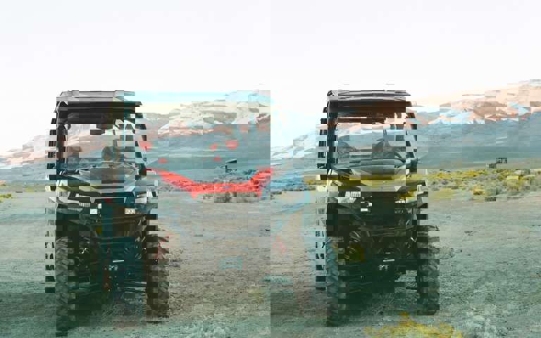 A red and black side-by-side vehicle parked in a dry landscape with sunlit mountain peaks in the distance.