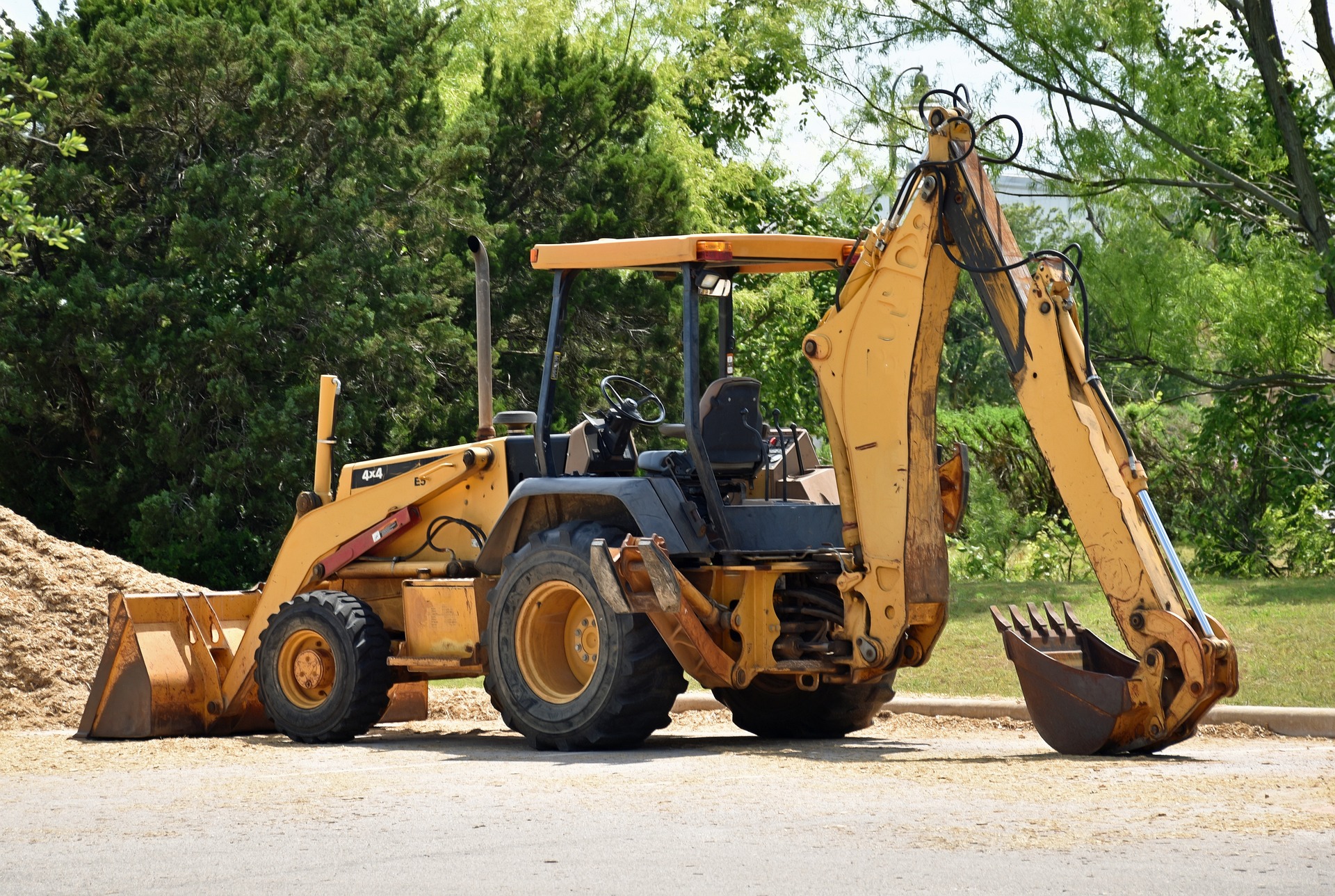 A yellow excavator positioned beside a dirt pile, set against a backdrop of green grass and trees.