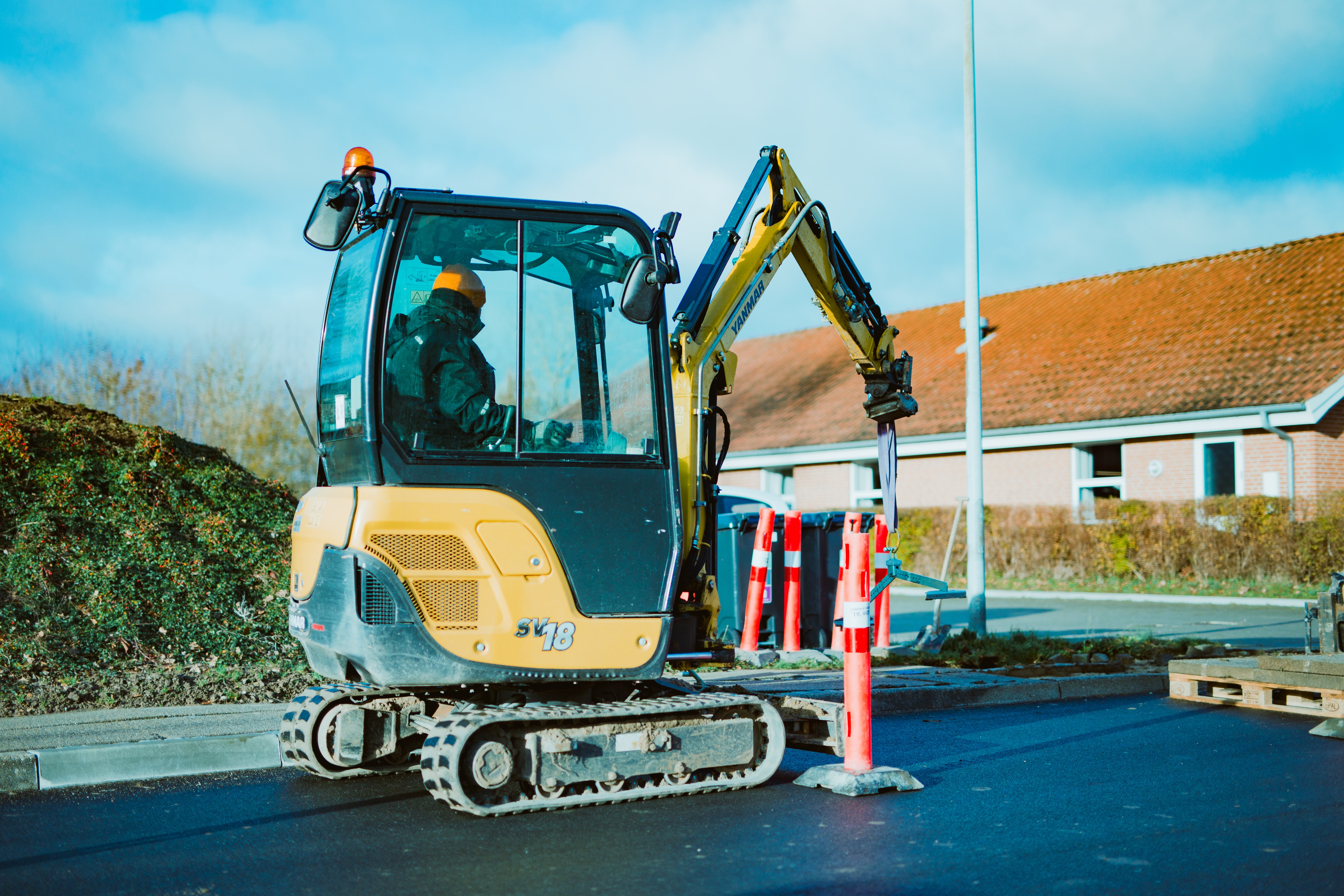 A worker uses a mini-excavator digger on a road, positioned in front of a single-story building.